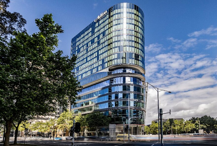 Glass tower hotel on a city corner with trees and blue sky.