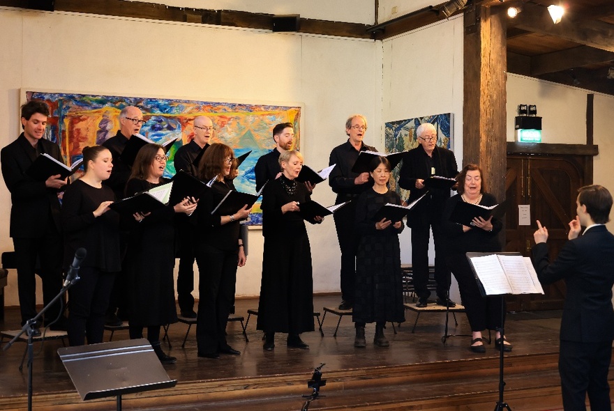 A choir of twelve adults all wearing black and singing from black songbooks, on a wooden stage with a colourful painting behind them.