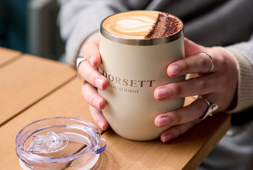 Two hands lighting resting on the outside of a cup of coffee on a wooden table.
