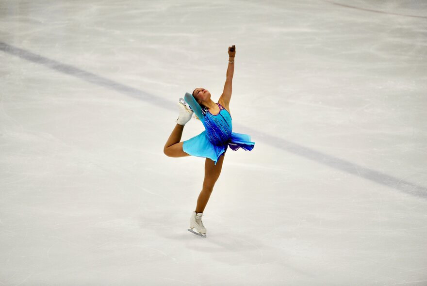 A figure skater on the ice rink wearing a blue costume holding a one-legged pose with their leg raised behind them.