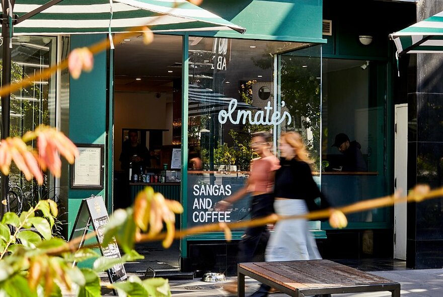 Exterior windows and entrance to Olmate sandwich shop with blurred pedestrians walking past, an outdoor table, and greenery in the foreground.