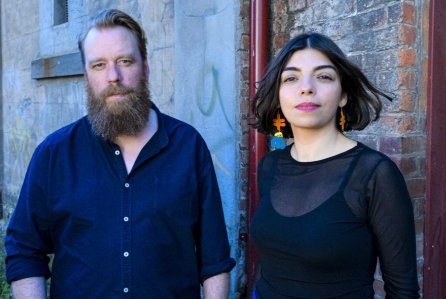A bearded man and a young woman standing side by side in front of a red brick building.