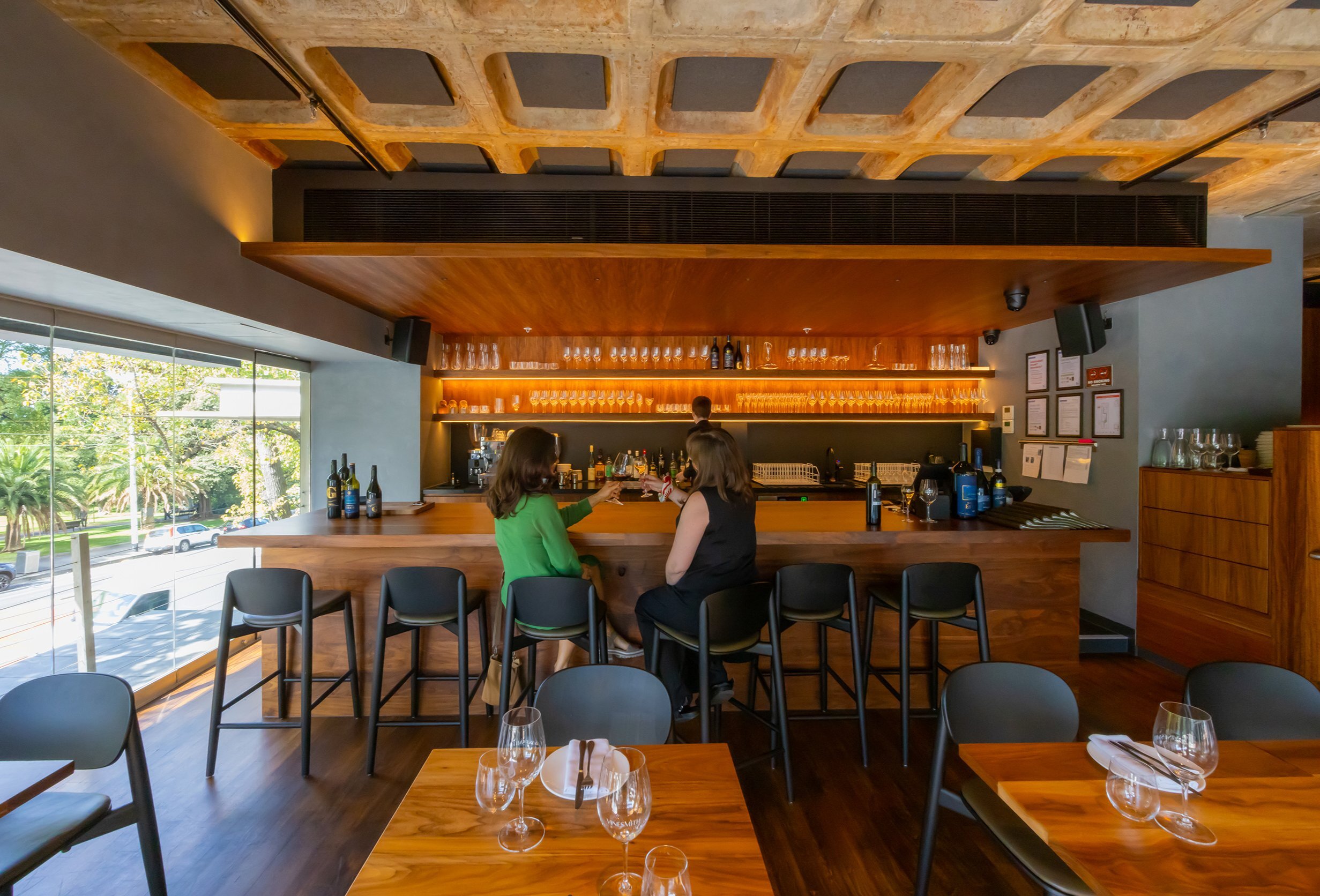 Two friends are doing a cheers at the bar of a warmly lit wine bar