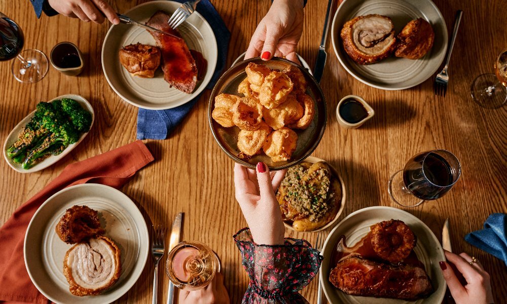 Someone passing a plate of Yorkshire puddings over a table set with rolled pork roast, veggies, wine and gravy.