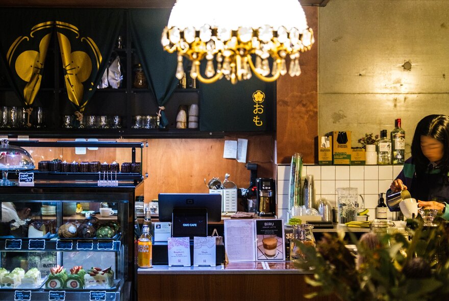 A barista is making coffee in a dimly-lit coffee shop. There is an ornate glass lamp hanging from the ceiling and a cabinet full of baked treats.