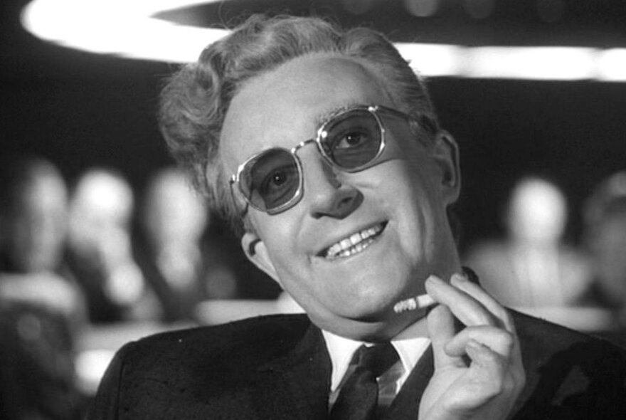 Peter Sellars smiling with a cigarette in hand in a black and white still image from the film Dr Strangelove.