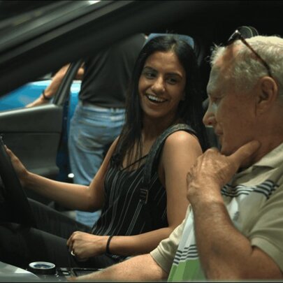 A young person smiling while sitting in the driver's seat of a car alongside an older passenger, at a motor show.