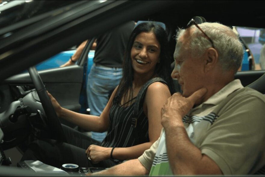 A young person smiling while sitting in the driver's seat of a car alongside an older passenger, at a motor show.