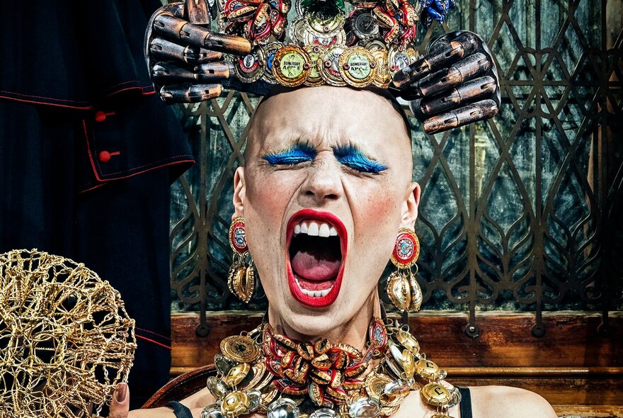 A screaming person with heavy makeup wearing an unusual headdress and necklace made from bottle caps and other scrap metal.