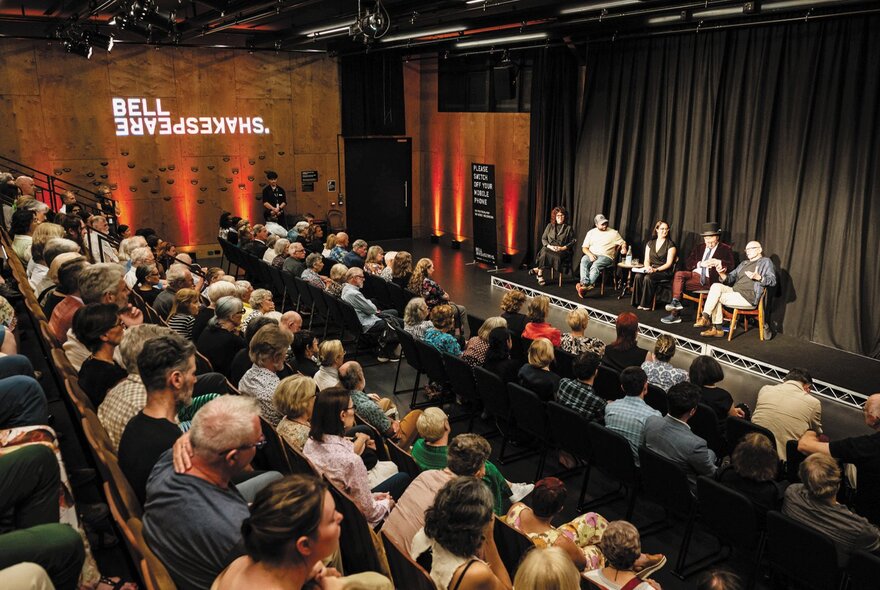 An audience seated in a lecture theatre for a panel discussion; five panellists on stage in front of a black stage curtain.