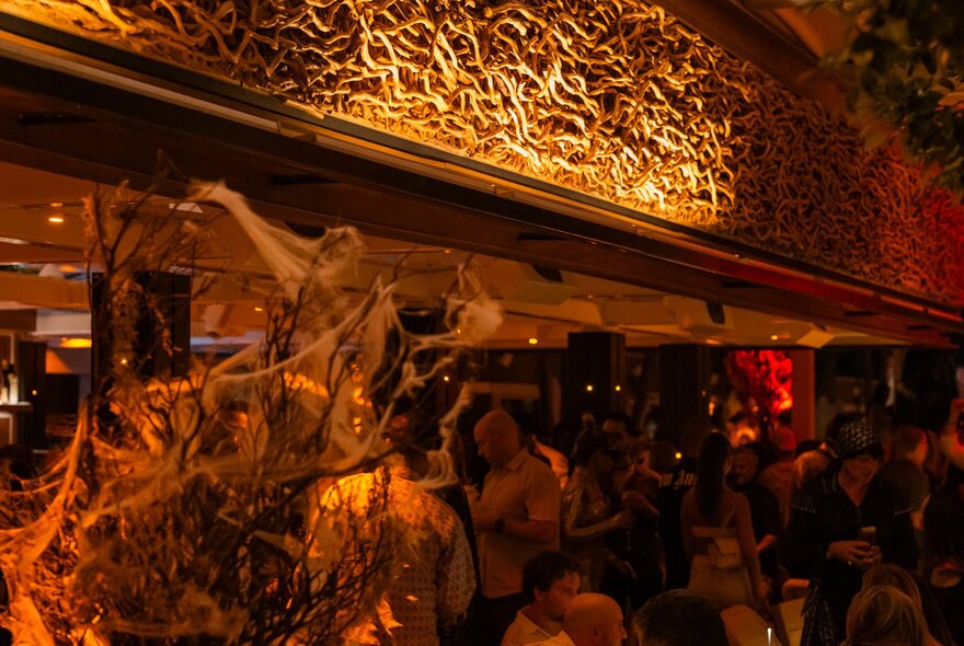 A dimly lit bar with patrons standing around and socialising, in the foreground is a spooky Halloween decoration of cobwebs on a tree branch.