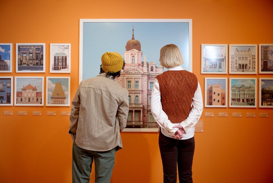 People standing in a gallery with orange walls looking at photos of buildings.