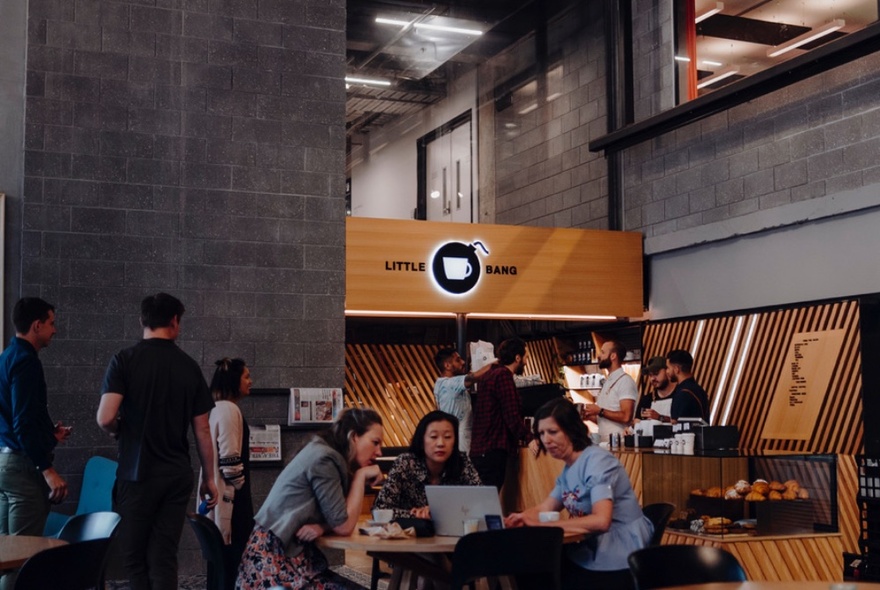 The interior of a busy cafe that has an industrial feel with grey concrete walls, and a wooden counter, with people lined up to order, and another group seated at a table and chairs.