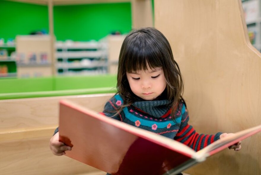 A young child reading a large red book at a library, with plywood shelving and green walls behind her.