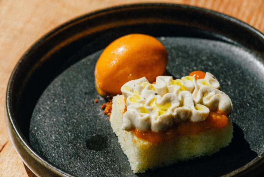 A square slice of an orange cake with a perfect dome of orange ice cream on a black plate.
