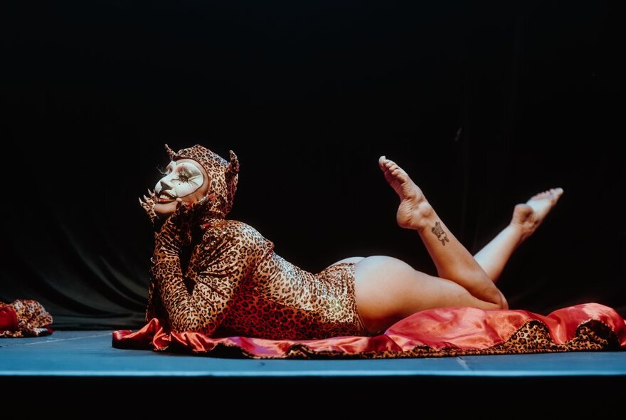 A performer wearing a cat or leopard costume lying on their stomach on a stage, their head and chest lifted and supported by their propped up elbow.