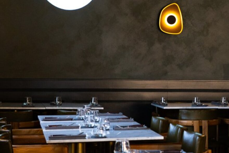 Interior of a dining room, with dark walls and a wall light, and tables set for service.