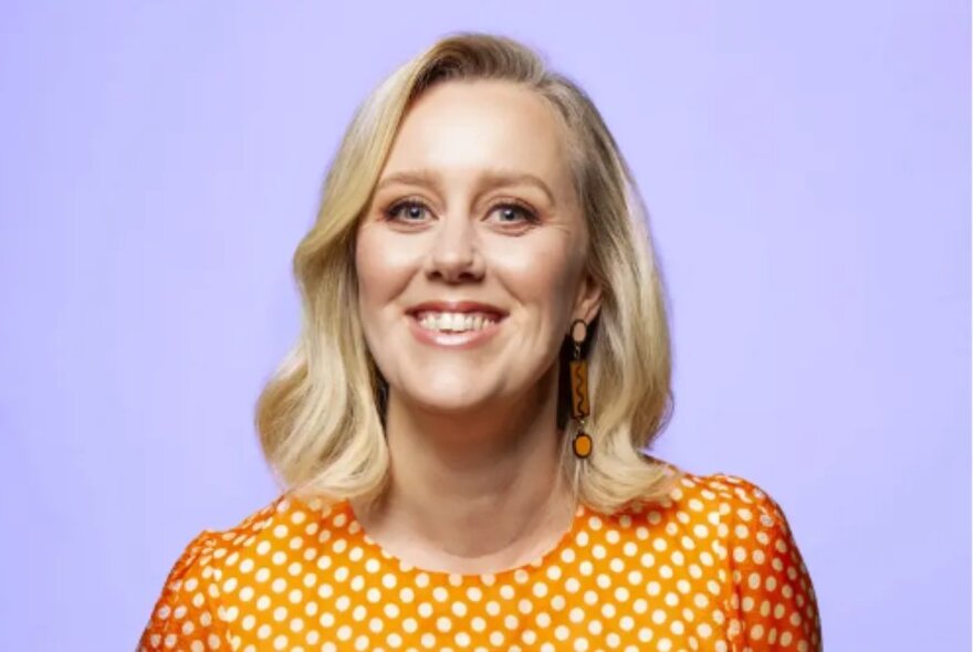 Blonde woman smiling wearing an orange polka-dote top against a light purple background.