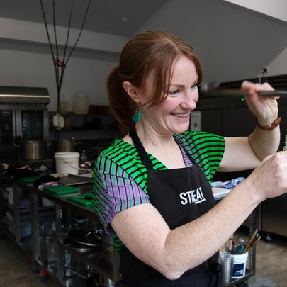 A woman in a green striped shirt and a black apron smiling while pouring a liquid into a jar from a ladle in a kitchen setting.