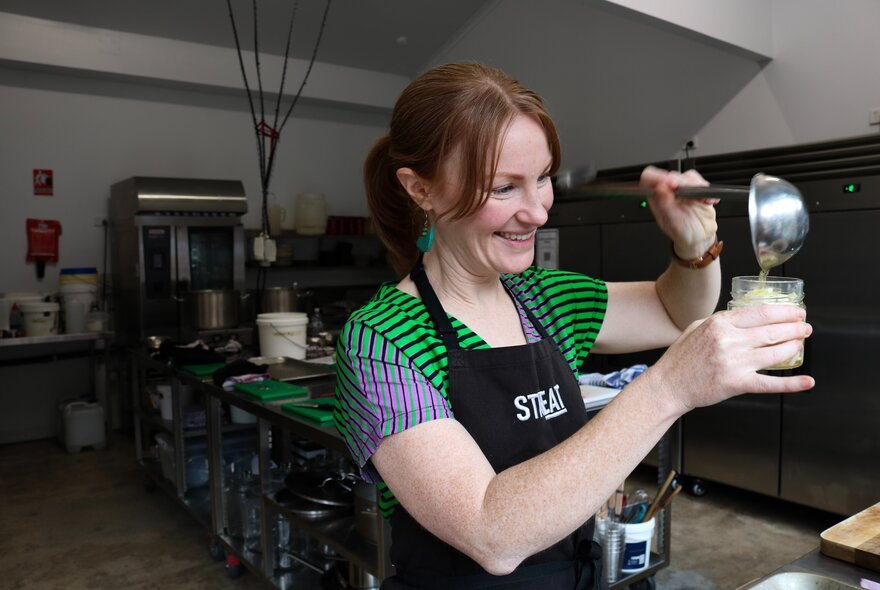 A woman in a green striped shirt and a black apron smiling while pouring a liquid into a jar from a ladle in a kitchen setting.