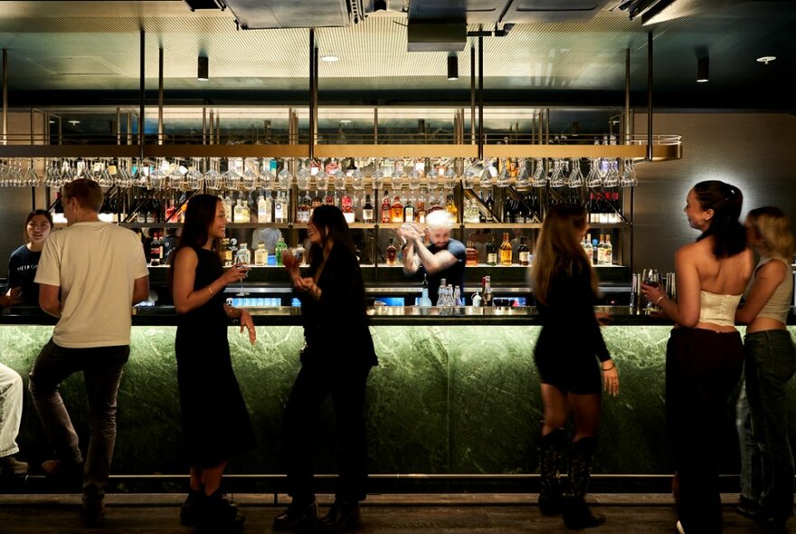 People standing lined up at a pub bar with undercounter lighting and shelves lined with bottles and glasses.