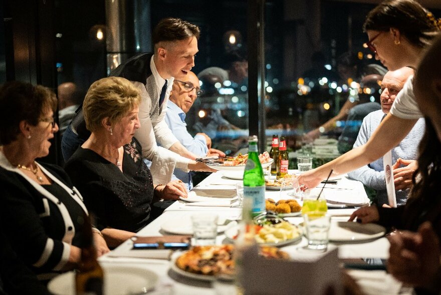 A long table inside a restaurant, with seated diners being served food by waiting staff. 