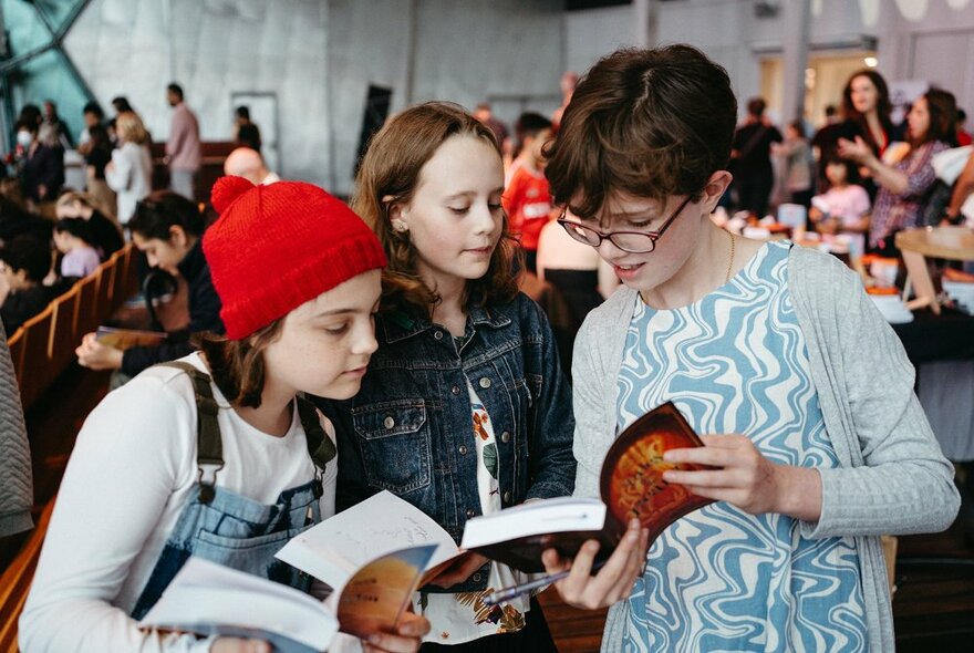 Three children comparing open books while a large group of children are seated in the background.