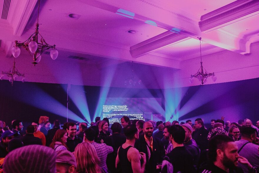 Interior of the Fringe Festival Hub venue, filled with people socialising and enjoying a drink, purple spotlights against a rear wall.