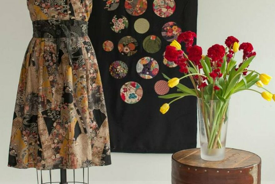 A hand-made Japanese print dress on a dressmakers' model, with a glass vase of red and yellow tulips beside and a black patchwork hanging behind.