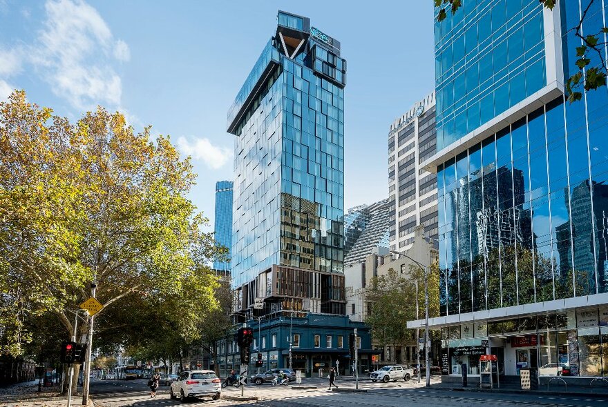 Exterior shot of glass-walled hotel tower building on street corner with trees and traffic.