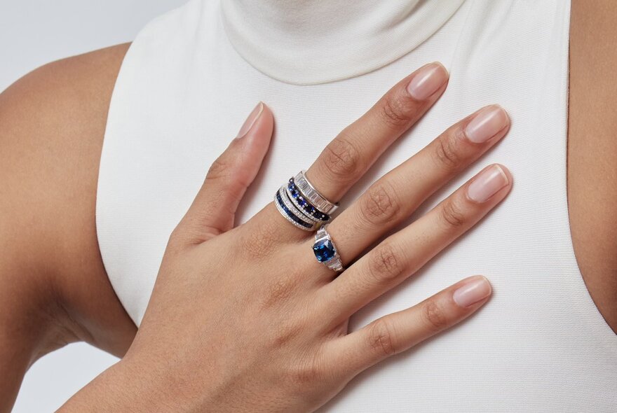 Model's ringed hand resting on her breastbone, wearing a sleeveless roll-neck white top.