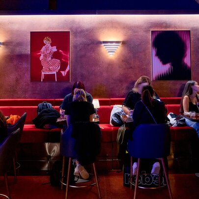People sitting in a moody restaurant with art on the walls and long red bench seating.