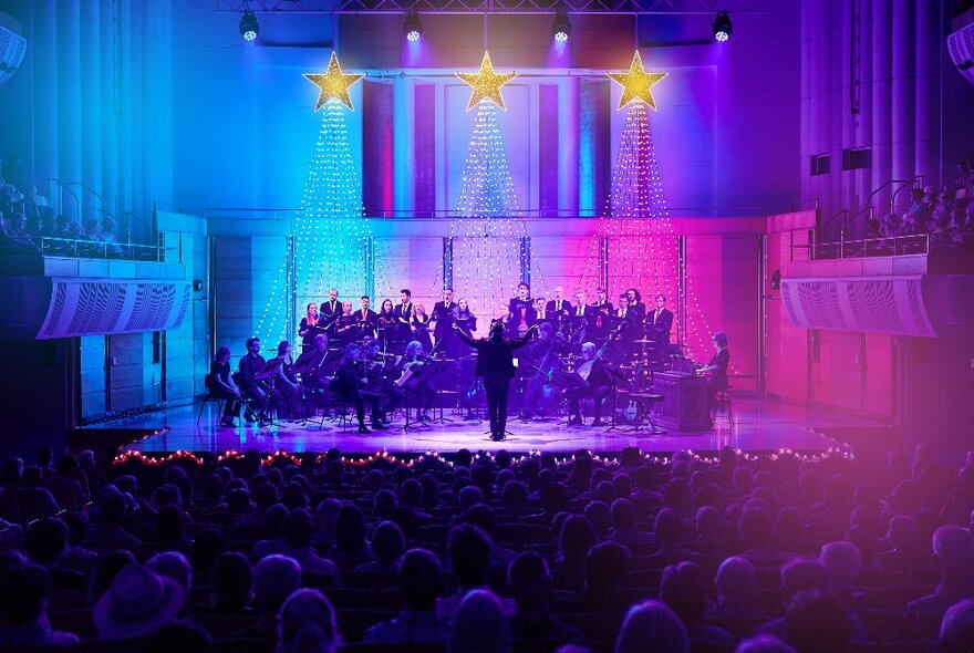 A massed choir and conductor on a large stage lit with blue, purple and pink lighting.