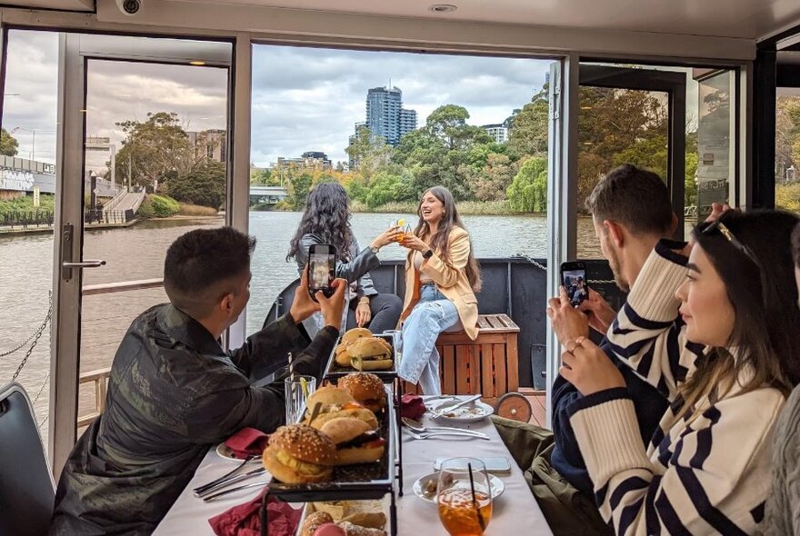 People seated inside a cabin of a riverboat, at a table with burgers and plates, taking photos of people seated outside on the rear of the boat.