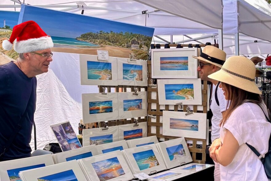 People wearing sunhats browsing seascape paintings at an outdoor market stall, the vendor wearing a Santa hat.