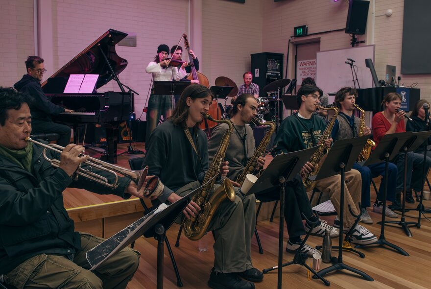 A room of musicians performing, with a horn section at the front and a grand piano at the back. 