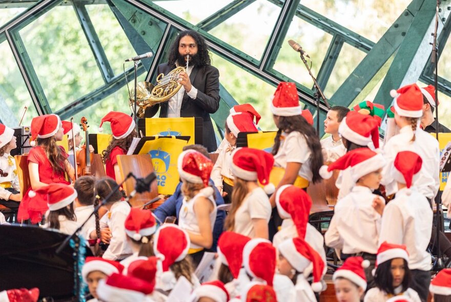 A choir of children wearing festive red Christmas hats looking at a musician playing a French horn.