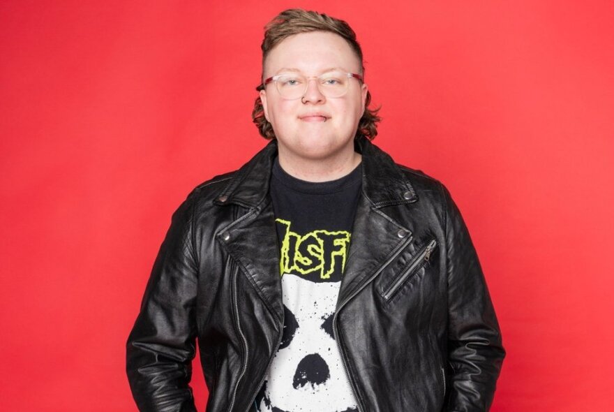 Comedian, Kaileb Hyland, wearing a black leather jacket and heavy metal t-shirt, smiling in front of a red background.