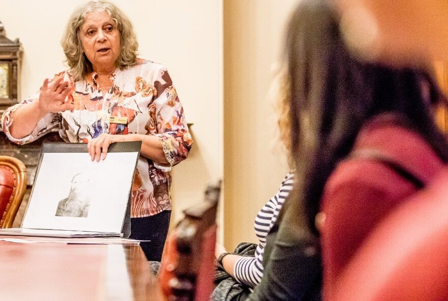 Woman with shoulder-length wavy hair, standing behind a table, indicating with right hand, holding a folder with sepia photograph, just visible, with left hand, talking to people on a tour.