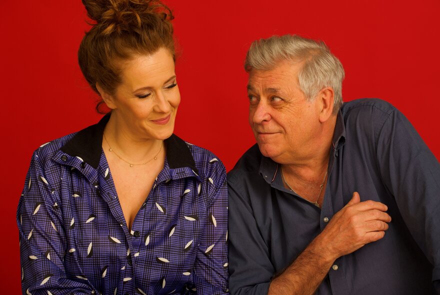 Italian musicians Riccardo Tesi and Maria Pierantoni Giua, looking at each other and posed against a red background.