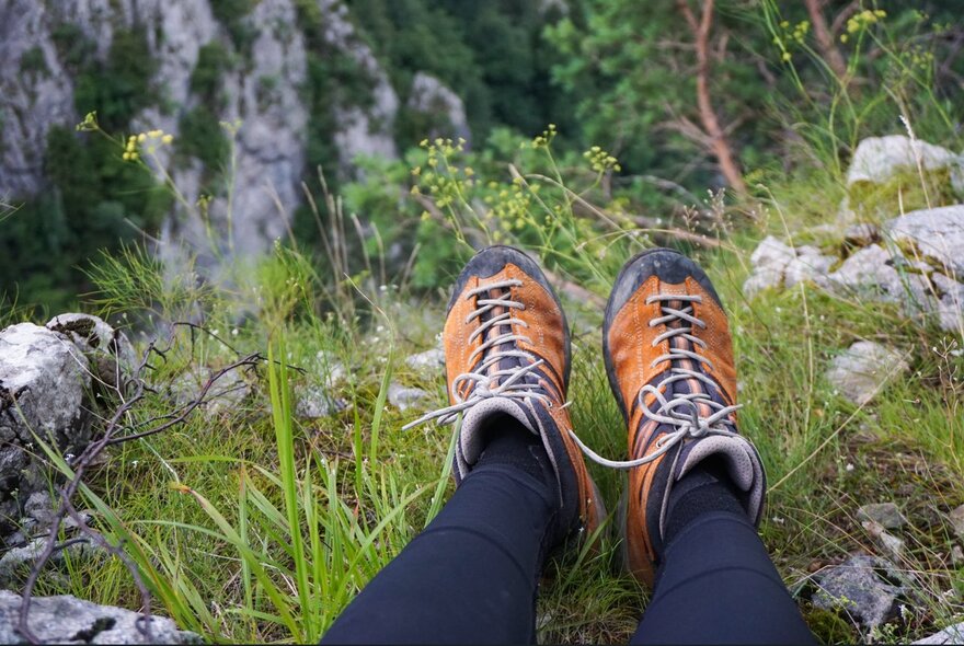 Lookinf down at legs with a pair of orange walking books in a grassy and rocky setting. 