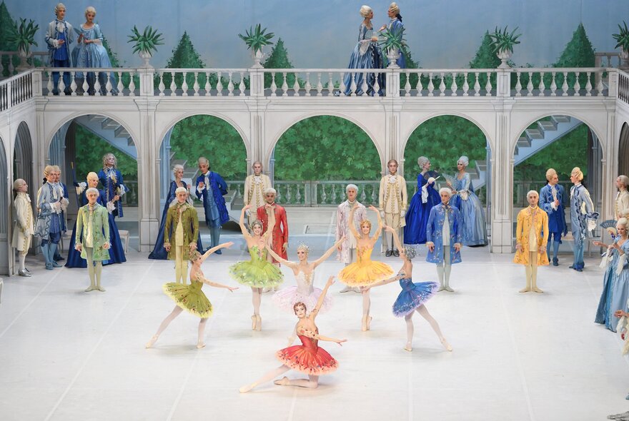 Ballet dancers in various coloured costumes performing on a stage with archways and a balcony. 