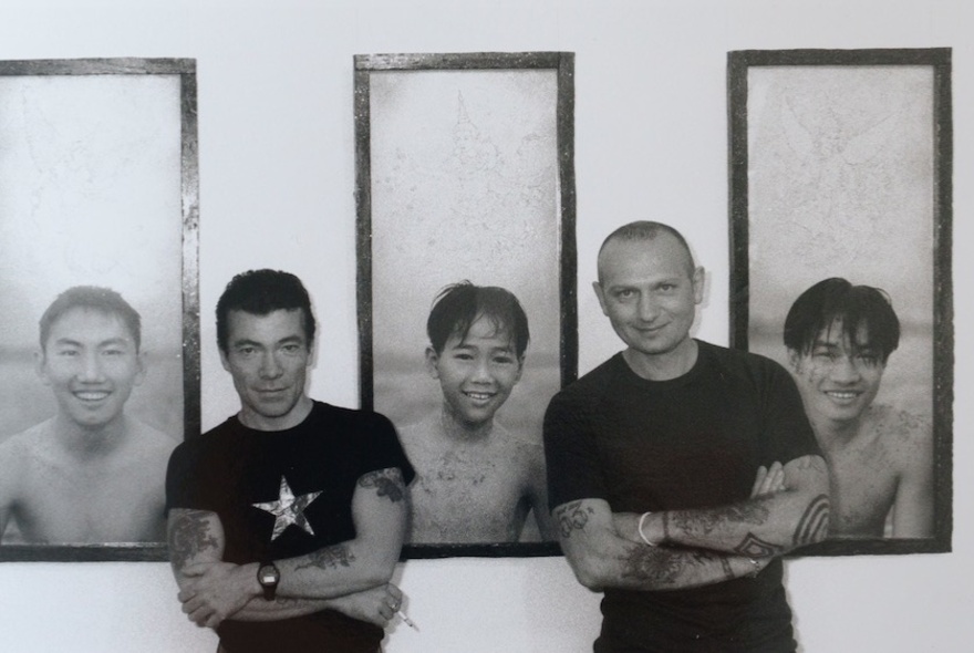 A black and white photograph of two male artists standing with their arms crossed in the gaps between three framed portraits of young Asian men on a wall.