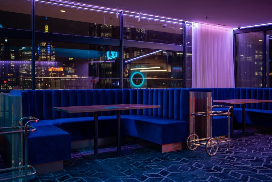 A retro neon interior of a bar with horseshoe banquettes and the city skyline in the background. 