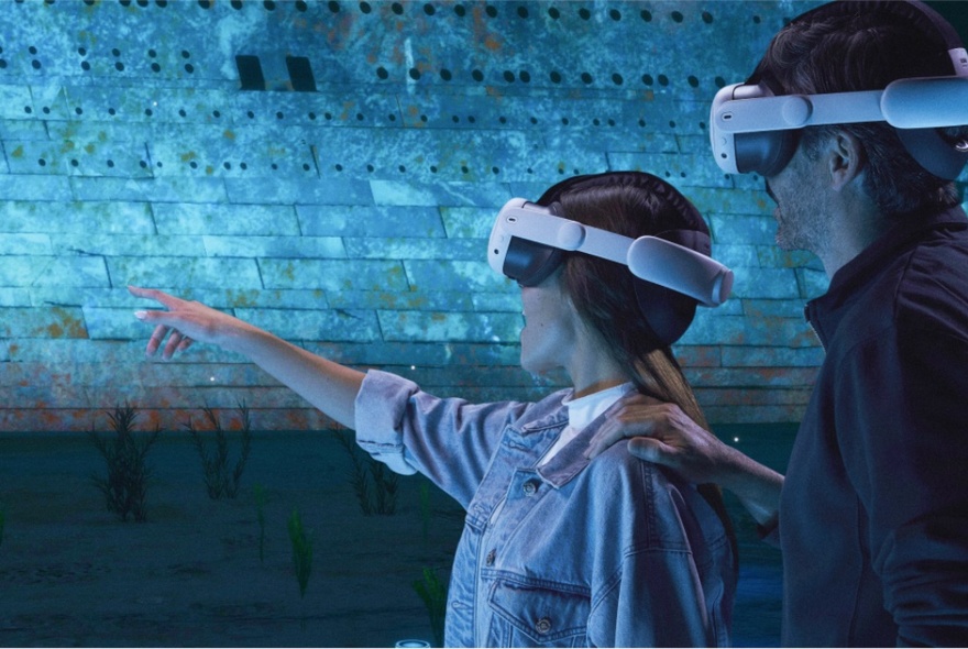 Two people in VR headsets in a darkened room, one pointing at something, the other with a hand placed on her shoulder. 