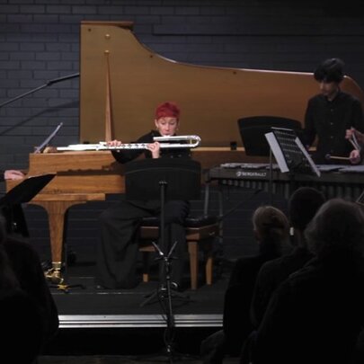 A live musical performance in front of a small audience featuring a flutist, a grand piano, a double bass, classical guitar and other instrumentalists.