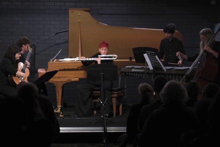 A live musical performance in front of a small audience featuring a flutist, a grand piano, a double bass, classical guitar and other instrumentalists.