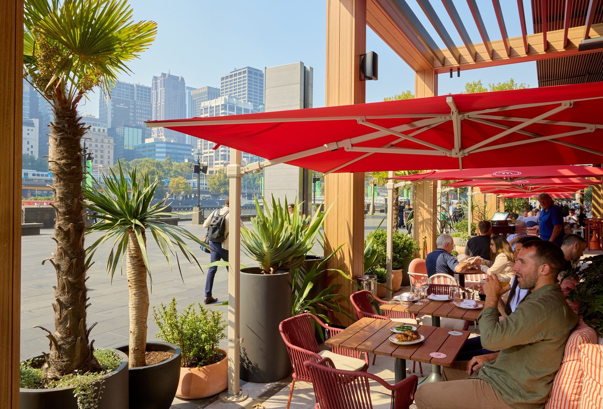 Two friends are drinking cocktails at a restaurant underneath a red sun umbrella. They are looking out onto a view of the city. 