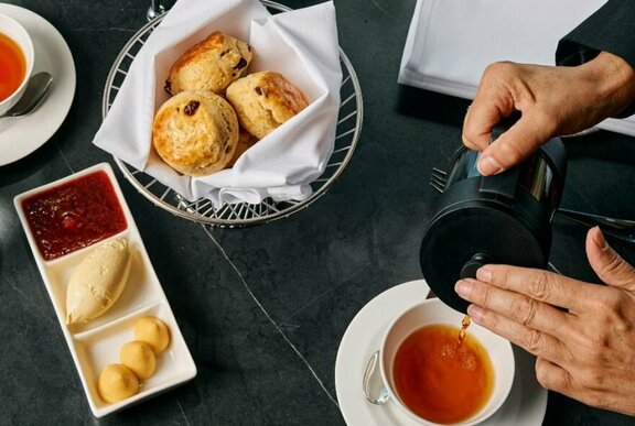 Afternoon tea spread with scones, jam and cream, hands pouring a cup of tea.