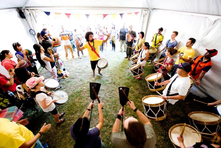 A percussion group in a large white tent with a leader in a yellow shirt standing in the middle. 